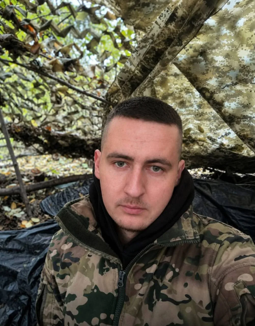 Портретне фото військового Володимира Пилипея на службі під час повномасштабної війни. Чоловік має коротке темне волосся й світлі очі. Він вдягнений в камуфляжну форму, стоїть на тлі маскувальної сітки. Фото з архіву сімʼї