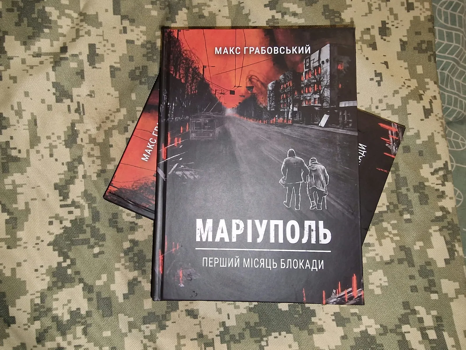Книга Макса Грабовського «Маріуполь. Перший місяць блокади» лежить на камуфляжній тканині поверх інших примірників. На обкладинці зображена зруйнована міська вулиця у червоно-чорних тонах.