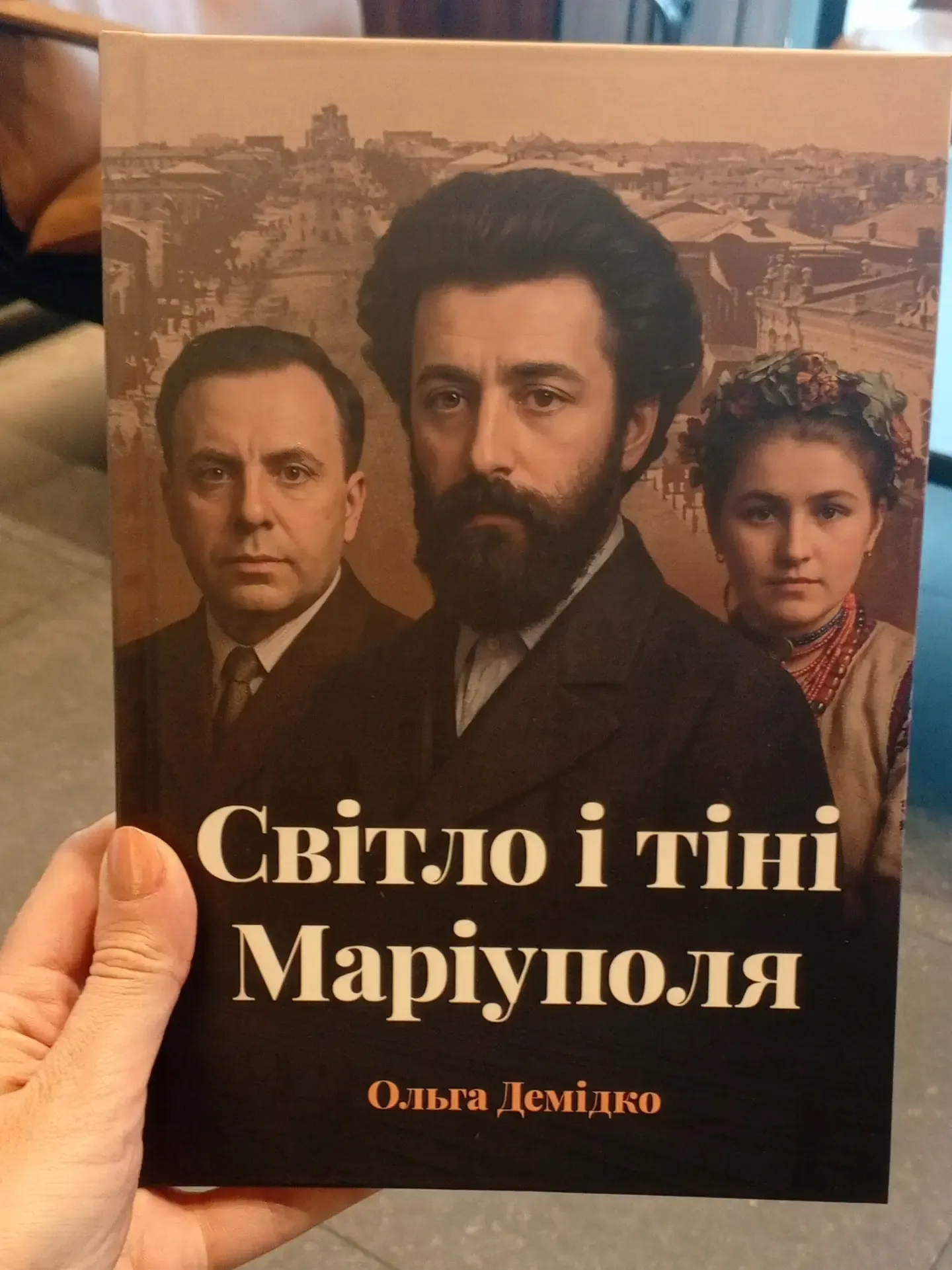 Обкладинка книги Ольги Демідко «Світло і тіні Маріуполя», яку тримають у руці. На обкладинці — портрети двох чоловіків і жінки в історичному вбранні на тлі старого міського пейзажу Маріуполя.
