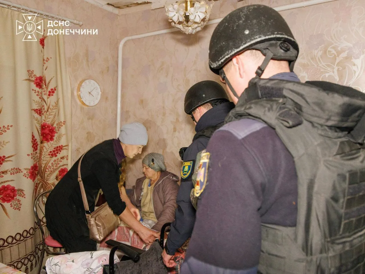 Рятувальники ДСНС у касках і бронежилетах допомагають жінкам під час евакуації з будинку. Одна жінка сидить на ліжку, інша нахилилася поруч із нею. Фото: ДСНС Донеччини