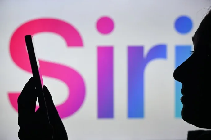 Зображення жінки, яка тримає мобільний телефон перед логотипом Siri, що відображається на екрані комп’ютера.