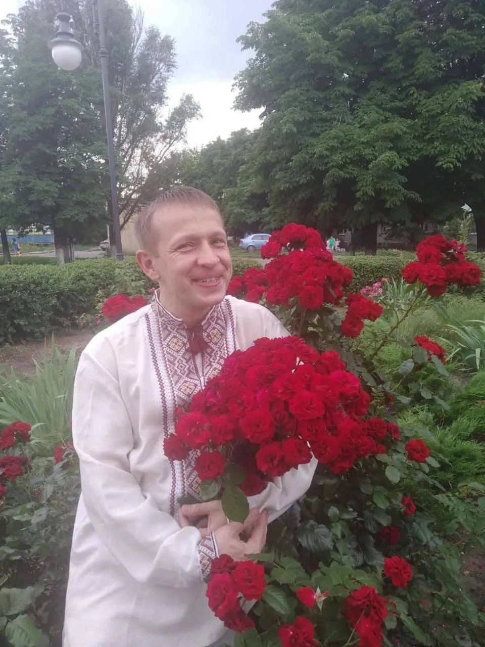 Чоловік у білій вишиванці усміхається та тримає великий букет червоних троянд у парку. Позаду видно дерева, клумби та вуличний ліхтар.