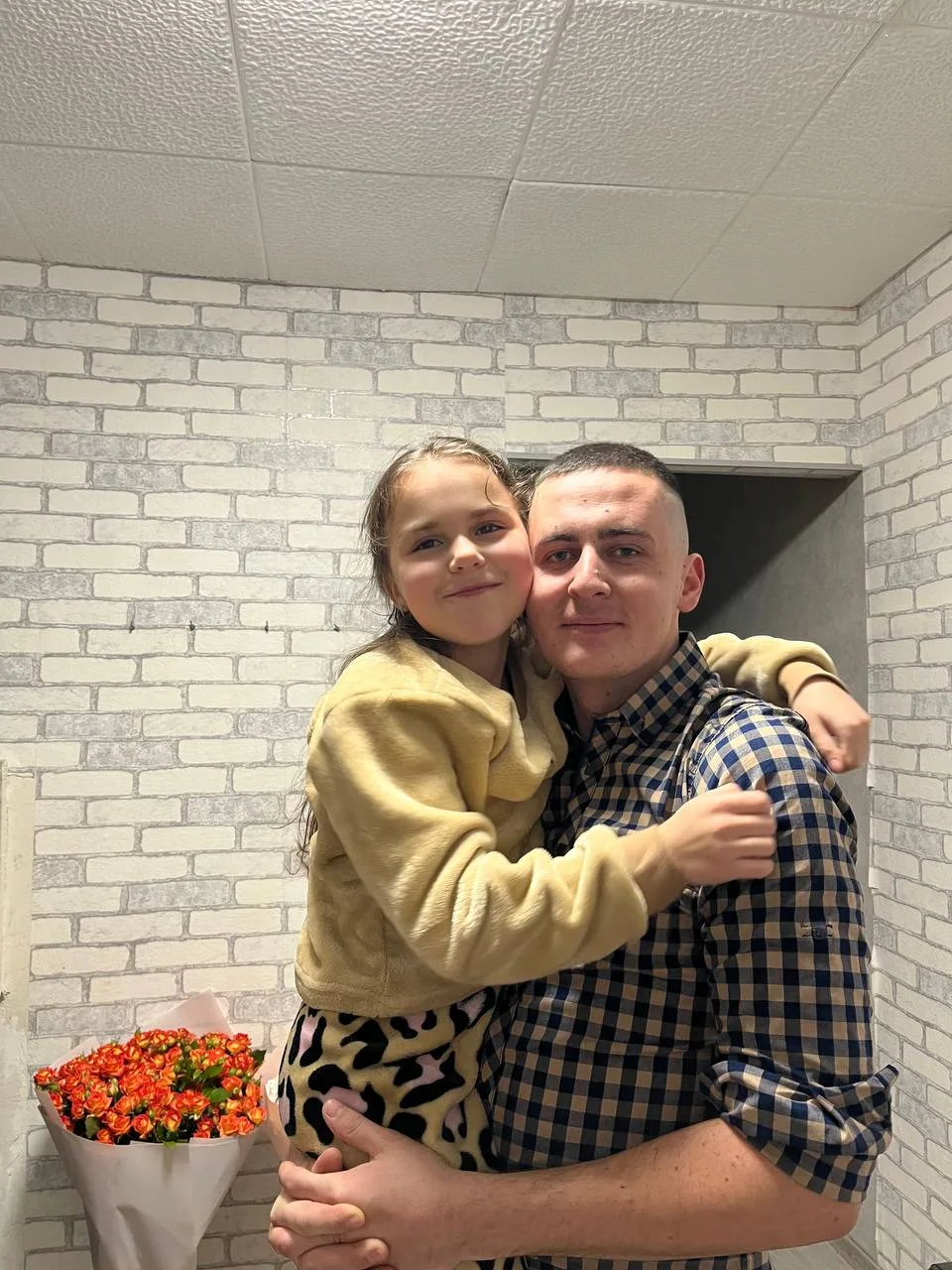 Військовий Володимир Пилипей із коротким темним волоссям й у картатій сорочці тримає на руках названу дочку Владиславу. Вона має довге темне волосся й вдягнена в жовтий світшот та світлі штани з плямистим візерунком. Дівчинка обіймає Володимира за плечі. Поруч на задньому плані — букет дрібних помаранчевих квітів. Фото з архіву сімʼї