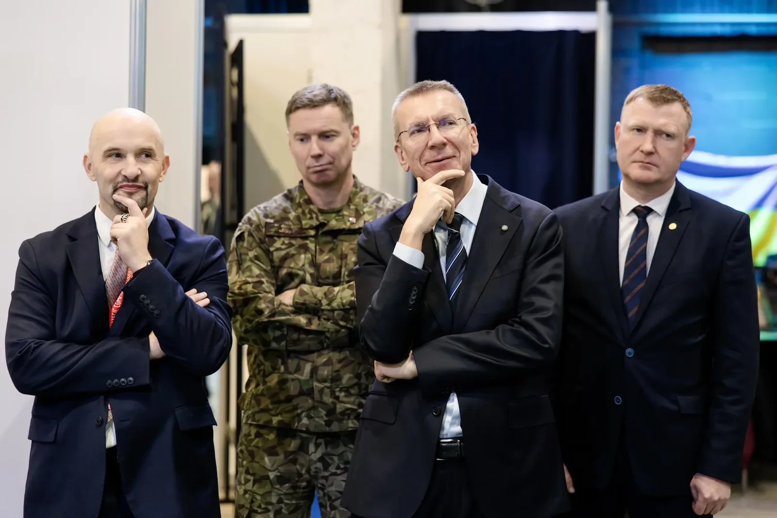 Четверо чоловіків стоять поруч: троє в темних костюмах, один у камуфляжній формі. Двоє чоловіків у костюмах торкаються підборіддя руками.
