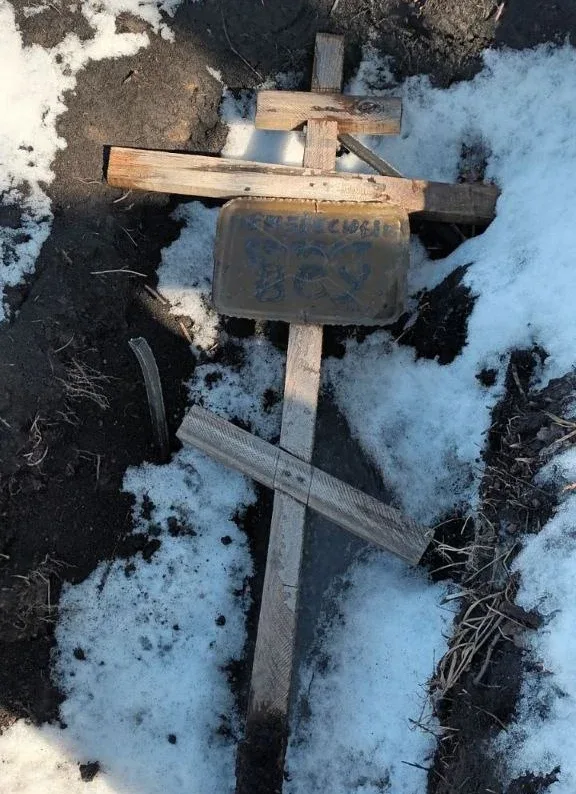 Дерев’яний хрест стоїть на землі серед залишків снігу. На перекладині закріплена табличка з написом.