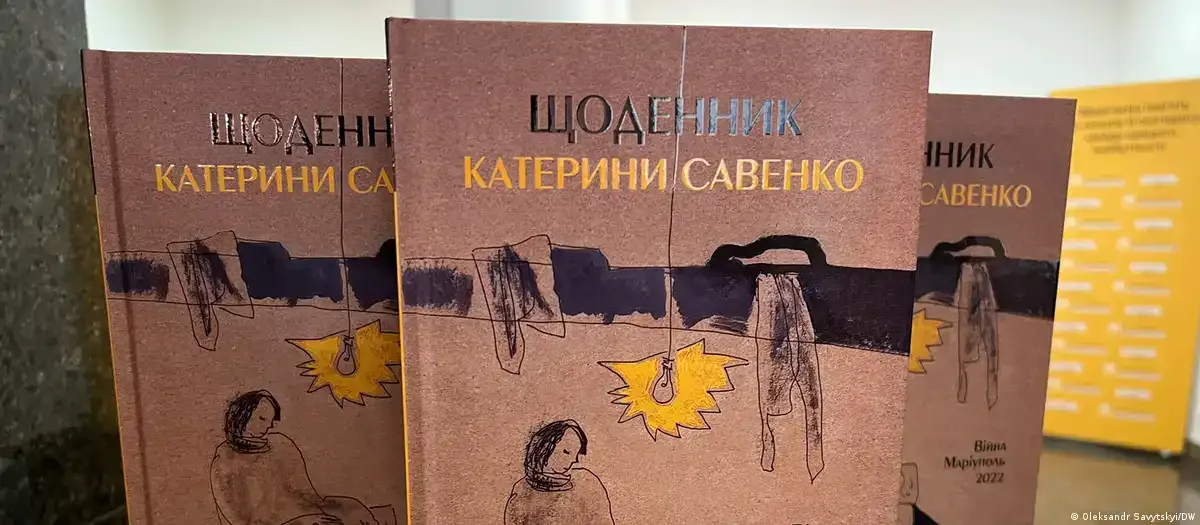 Три примірники книги «Щоденник Катерини Савенко» стоять поруч. На обкладинці — малюнок із людиною, речами на мотузці та лампою на коричневому тлі.