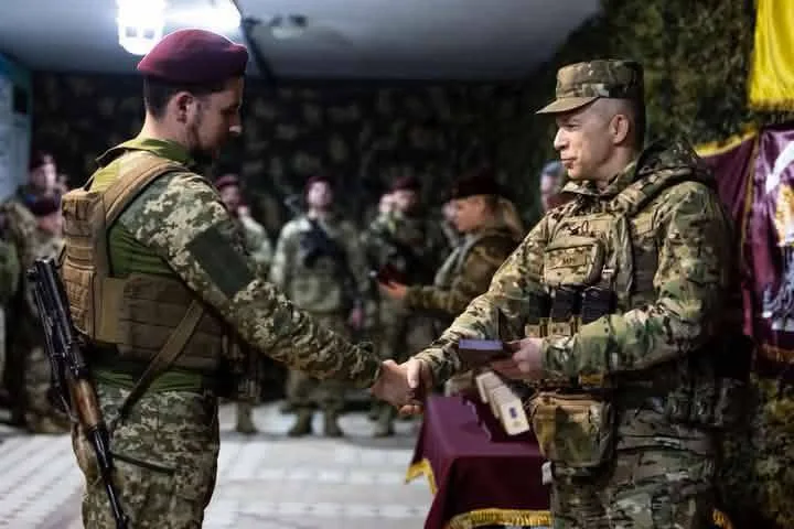 Військовий Володимир Пилипей у формі з піксельним камуфляжем та в бордовому береті тисне руку тодішньому Головнокомандувачу Збройних сил України Олександру Сирському. В одній руці вдягнений по форму Сирський тримає нагороду. На задньому плані стоять інші військовослужбовці. Фото з архіву сімʼї