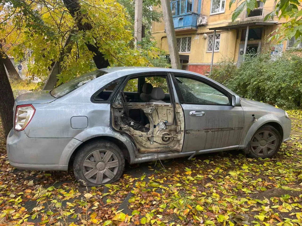 Пошкоджений сірий легковий автомобіль стоїть у дворі біля багатоповерхового будинку. На машині відсутня обшивка задніх лівих дверей, вибите скло та видно пошкодження кузова.