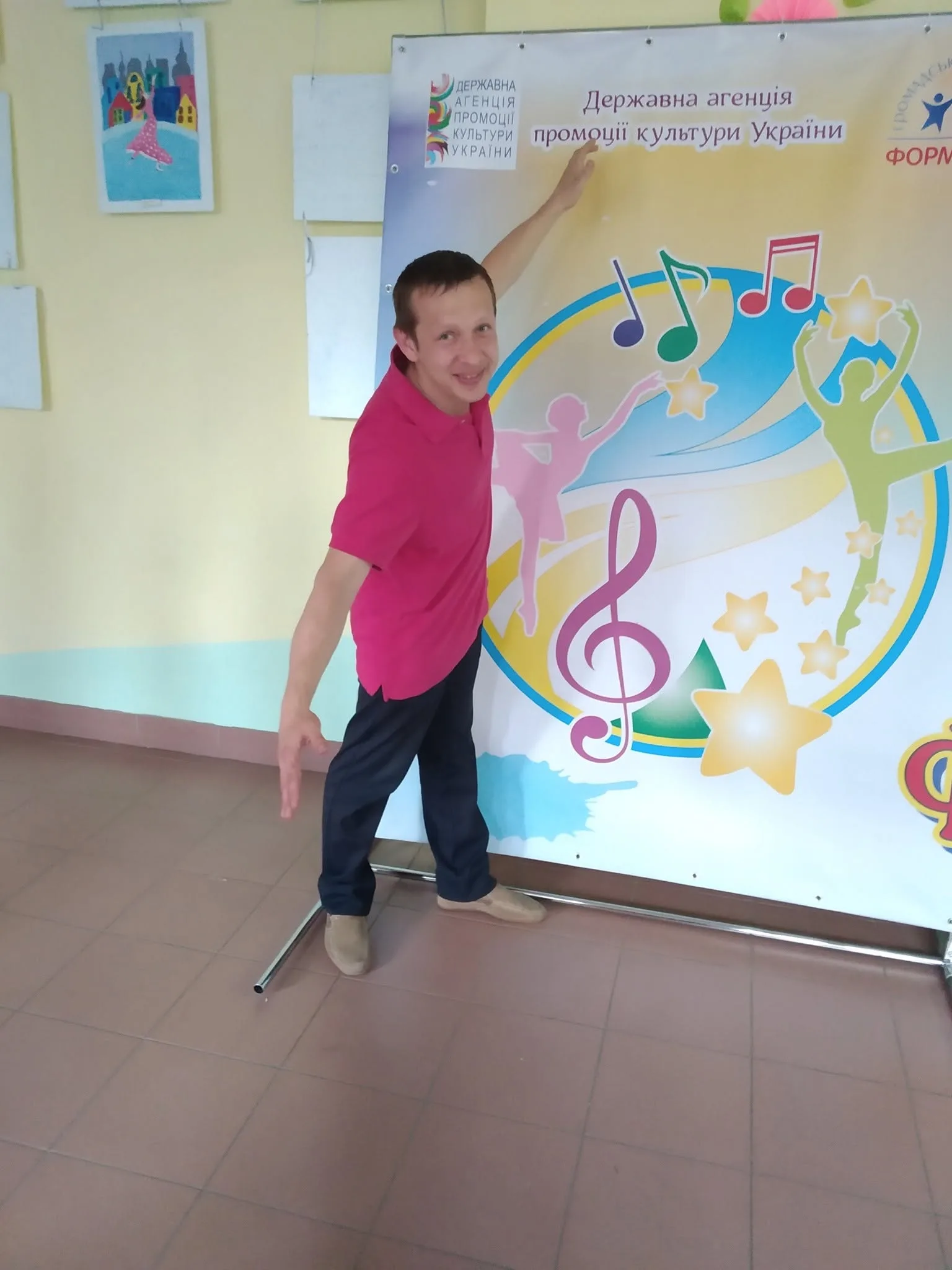 Чоловік у рожевій футболці позує біля банера Державної агенції промоції культури України з музичними символами та силуетами танцівників. На стіні поруч видно дитячі малюнки.