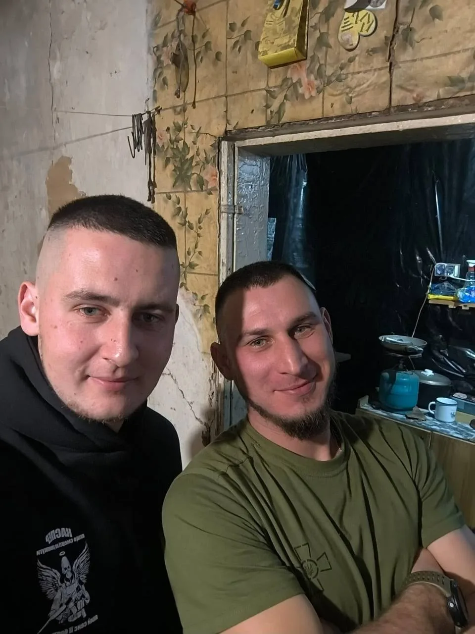 Військовий Володимир Пилипей із другом і побратимом Романом: Володимир ліворуч — із коротким темним волоссям, світлими очима та короткою щетиною, у чорному худі з білим графічним принтом; Роман праворуч — із коротким темним волоссям, світлими очима та густою темною борідкою, у зеленій футболці й наручному годиннику. Чоловіки фотографуються у приміщенні з обшитими плиткою стінами та облаштованою кухонною зоною на задньому плані. Фото з архіву сімʼї