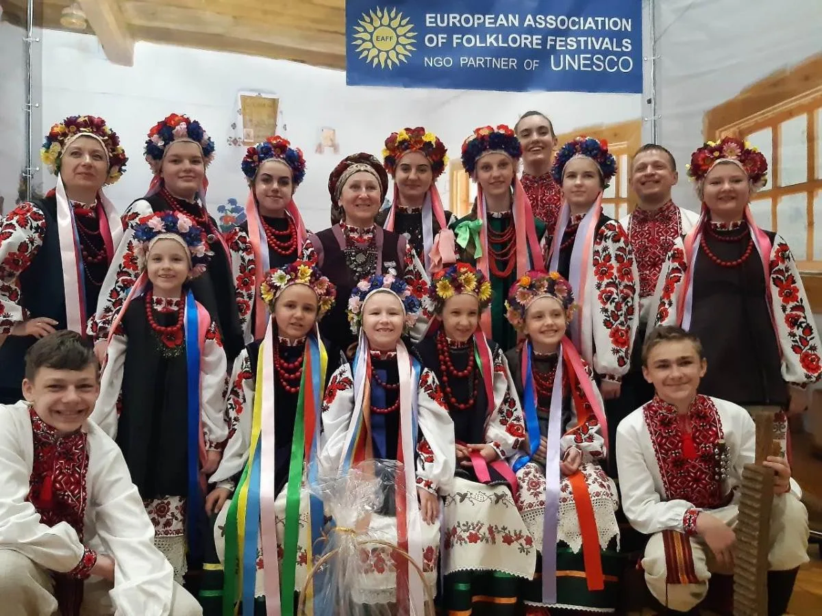Групове фото дітей і дорослих у традиційному українському вбранні та квіткових вінках. Учасники позують під банером Європейської асоціації фольклорних фестивалів — партнера ЮНЕСКО
