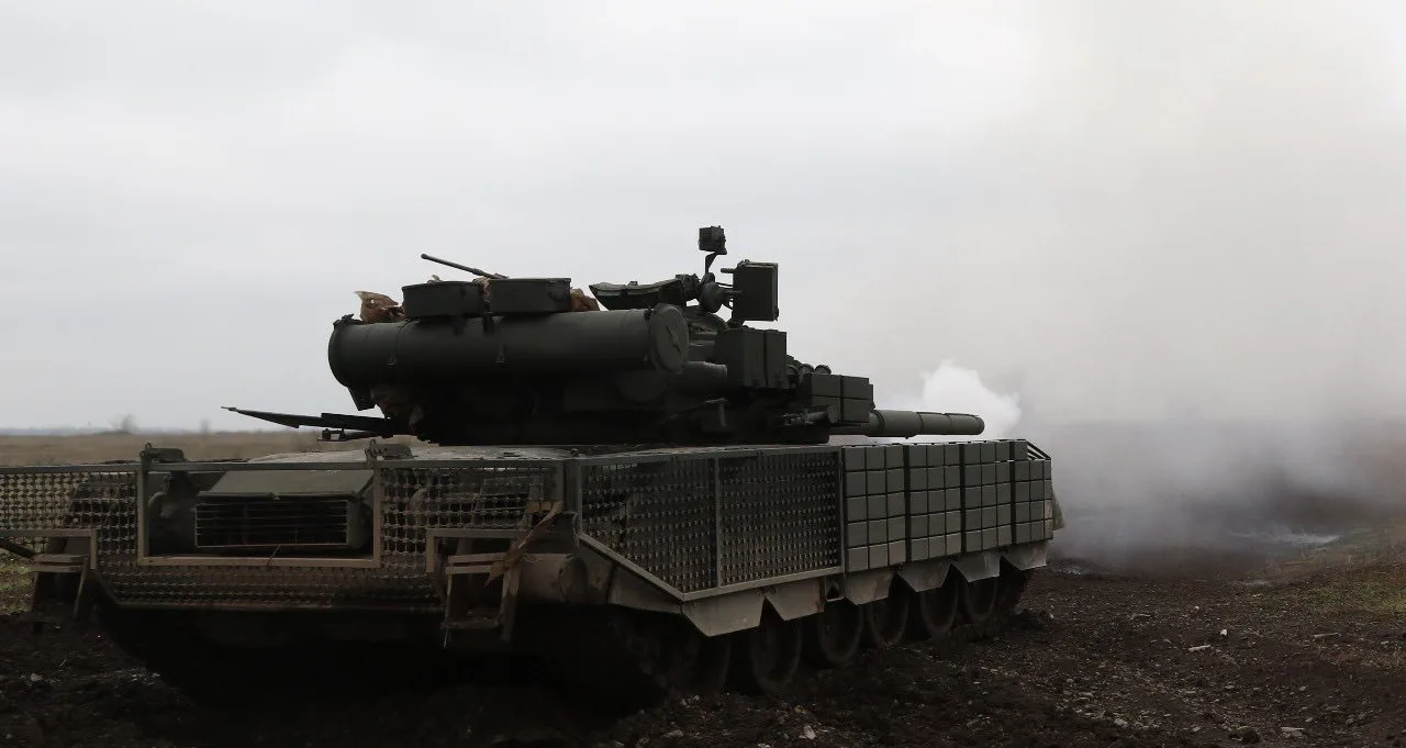 Танк із додатковим захистом на корпусі, який рухається по розмитому польовому ґрунту в туманну погоду. Зі ствола підіймається дим, що свідчить про нещодавній постріл або роботу двигуна під час бойових дій.