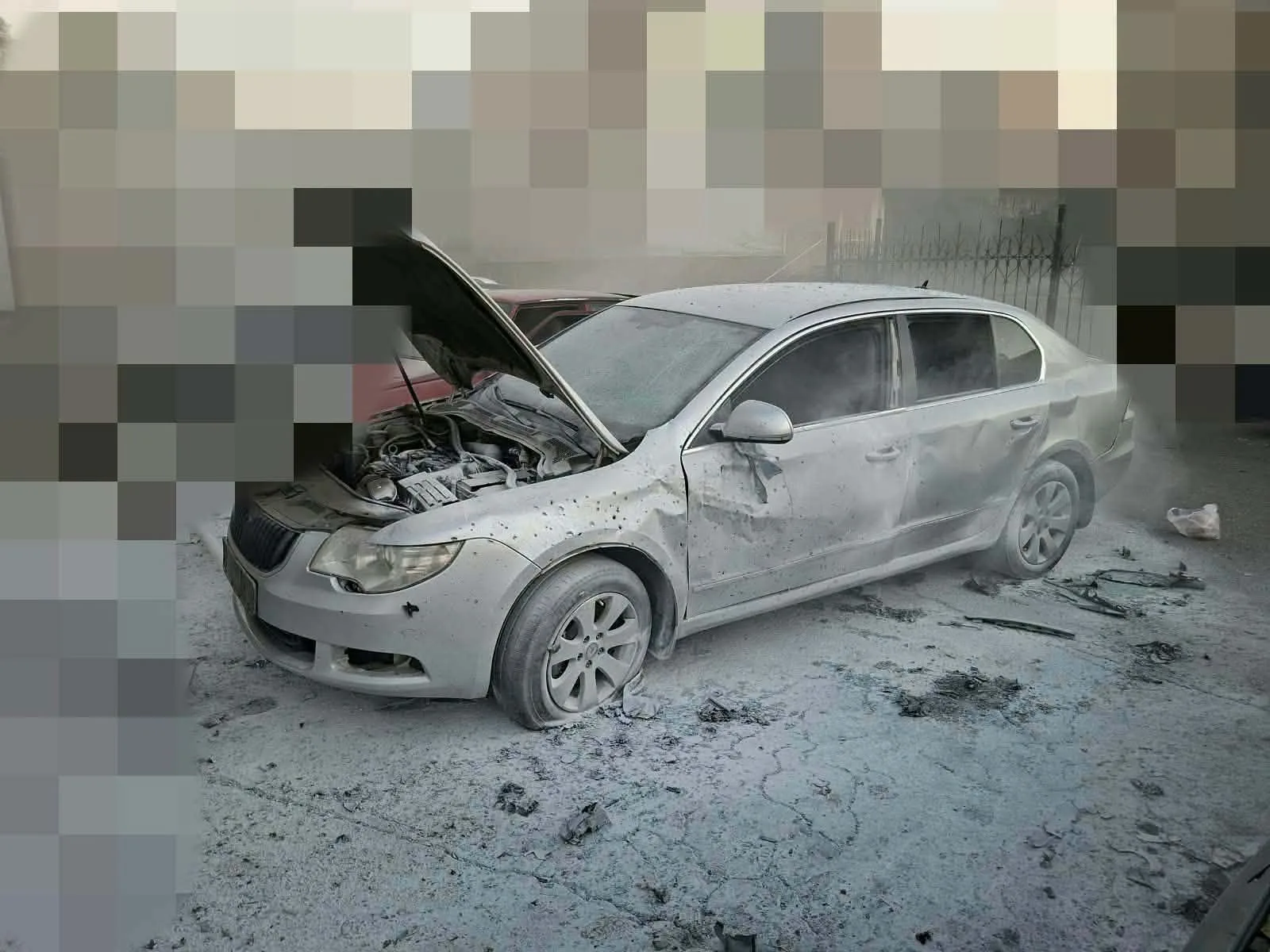 Пошкоджений сріблястий легковий автомобіль із відкритим капотом стоїть серед уламків на дорозі. На кузові видно вм’ятини та сліди пошкоджень, довкола — уламки й пил, тло зображення частково пікселізоване