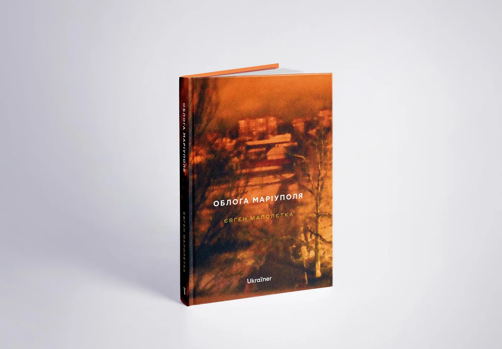 Книга Євгена Малолєтки «Облога Маріуполя» стоїть на світлому тлі. На обкладинці — розмите фото міста в помаранчево-чорних тонах