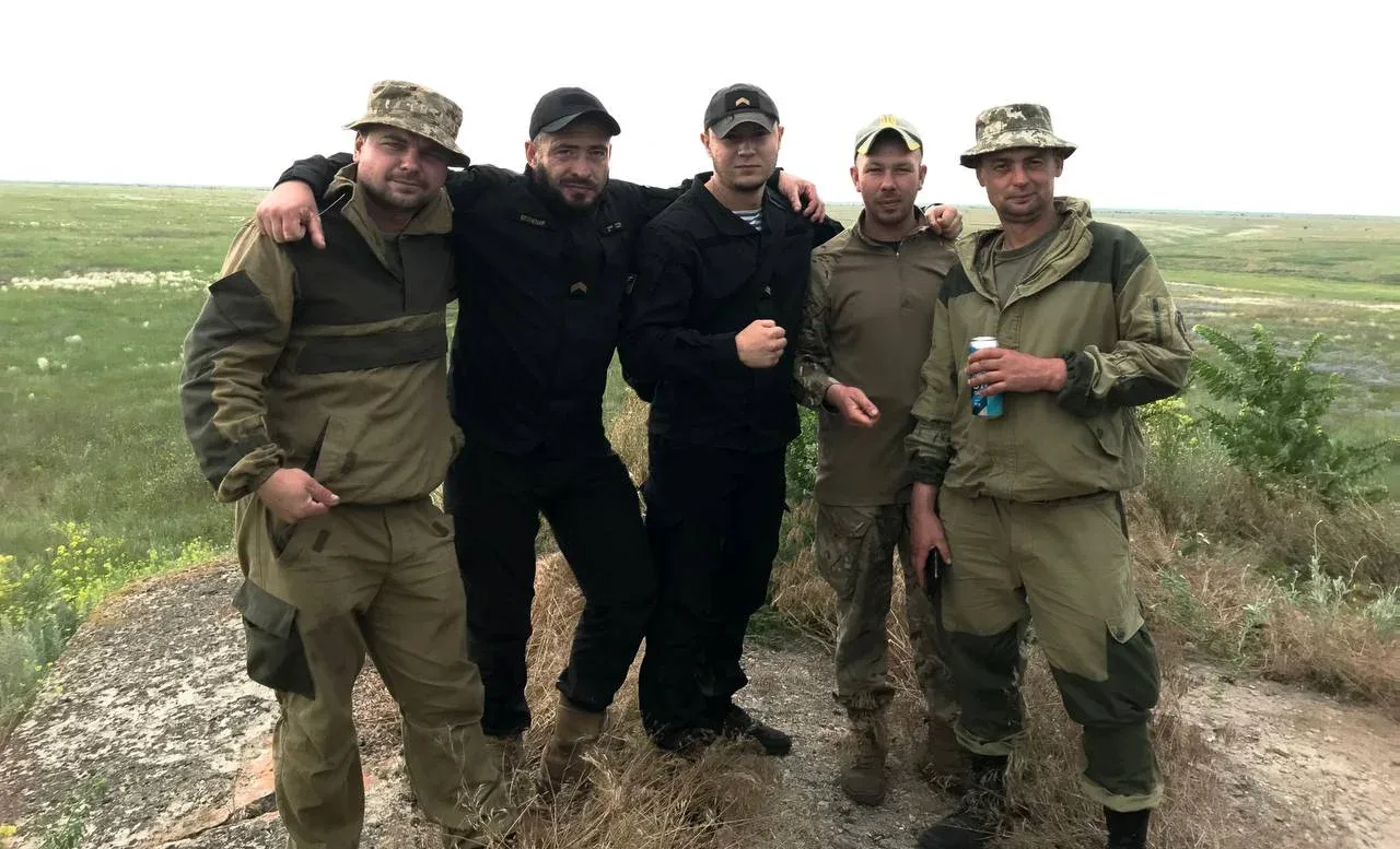П’ятеро чоловіків стоять разом на відкритій місцевості. Двоє обіймають інших за плечі, а чоловік праворуч тримає бляшанку.