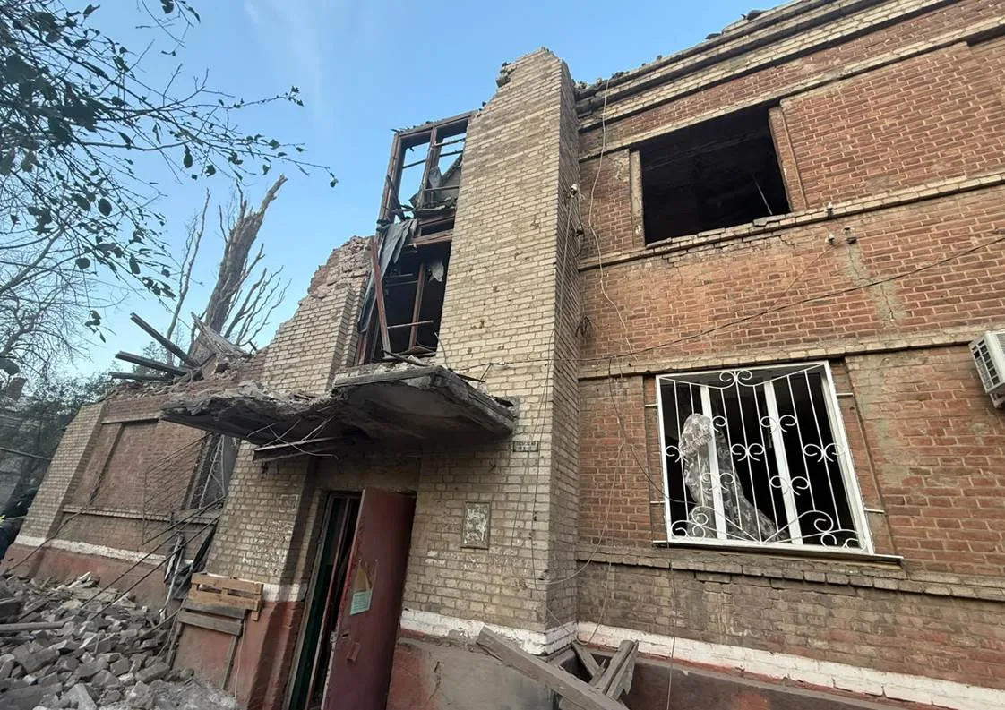 Пошкоджена цегляна будівля з частково обваленими стінами та перекриттями. У вікнах вибите скло, частина даху зруйнована, біля входу та вздовж фасаду лежать уламки цегли й будівельних конструкцій.