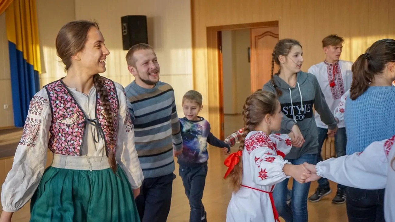 Група дітей і дорослих тримається за руки під час спільного заняття в приміщенні. Частина учасників одягнена у вишиванки, ліворуч стоїть усміхнена жінка в традиційному вбранні, поруч — чоловік у смугастому светрі.