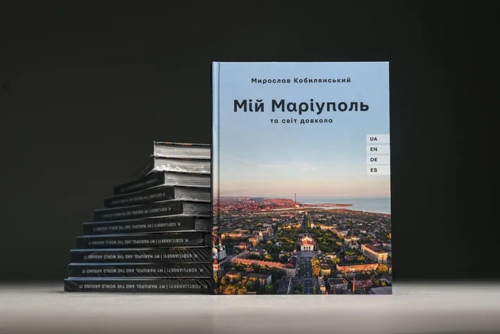 Книга Мирослава Кобилянського «Мій Маріуполь та світ довкола» стоїть на столі поруч зі стосом інших примірників. На обкладинці — панорамний вид Маріуполя з міською забудовою та Азовським морем удалині