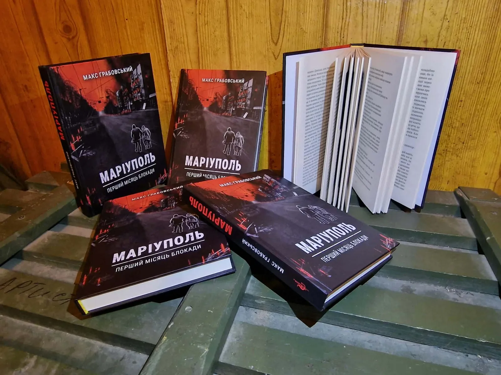 Кілька примірників книги Макса Грабовського «Маріуполь. Перший місяць блокади» лежать на столі, один примірник відкритий. На обкладинках — зображення зруйнованого міста у червоно-чорних тонах.