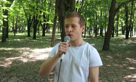 Чоловік у світлій футболці тримає мікрофон і виступає просто неба в зеленому парку. На задньому плані — дерева та затінена галявина.
