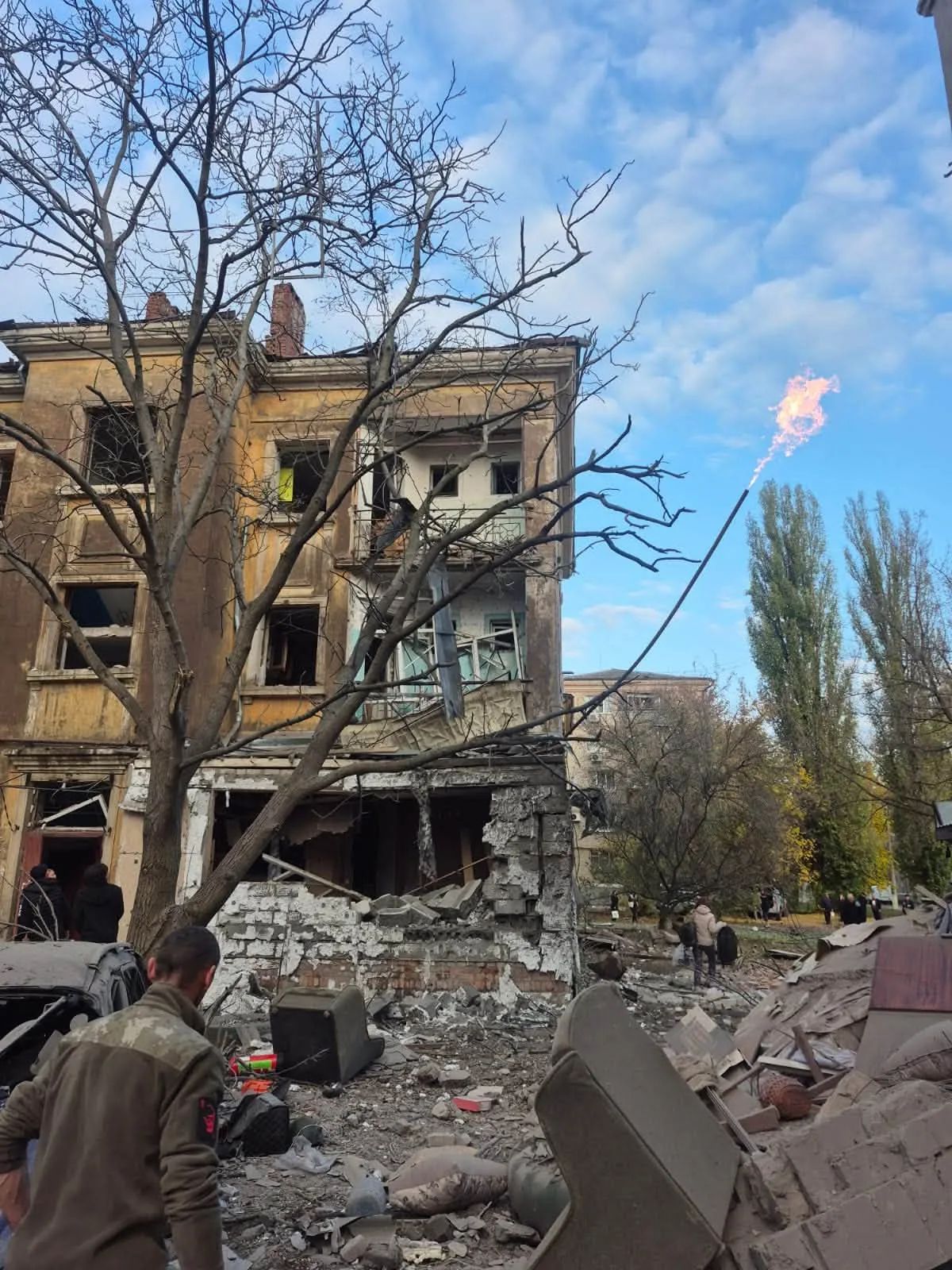 Зруйнований багатоповерховий будинок із вибитими вікнами та пошкодженим фасадом. Перед будівлею лежать уламки конструкцій і частини майна, поруч перебувають люди. Праворуч видно пошкоджену газову трубу, з якої виривається полум’я.