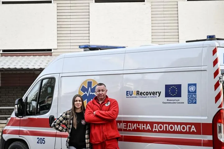 Двоє працівників екстреної медичної допомоги Запоріжжя стоять біля автомобіля з логотипами EU4Recovery, ЄС та ПРООН.