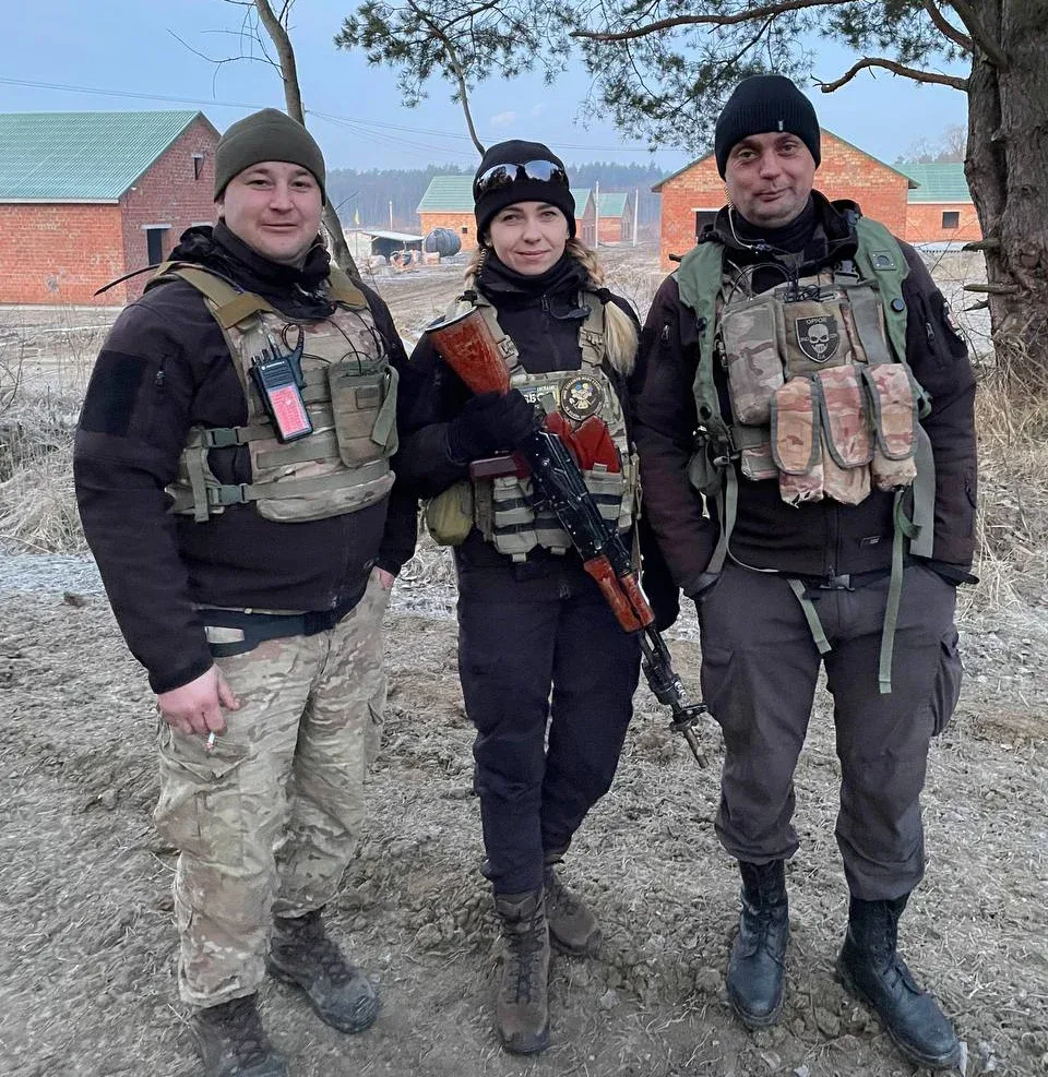 Двоє чоловіків і жінка стоять поруч у захисних жилетах. Жінка посередині тримає зброю.