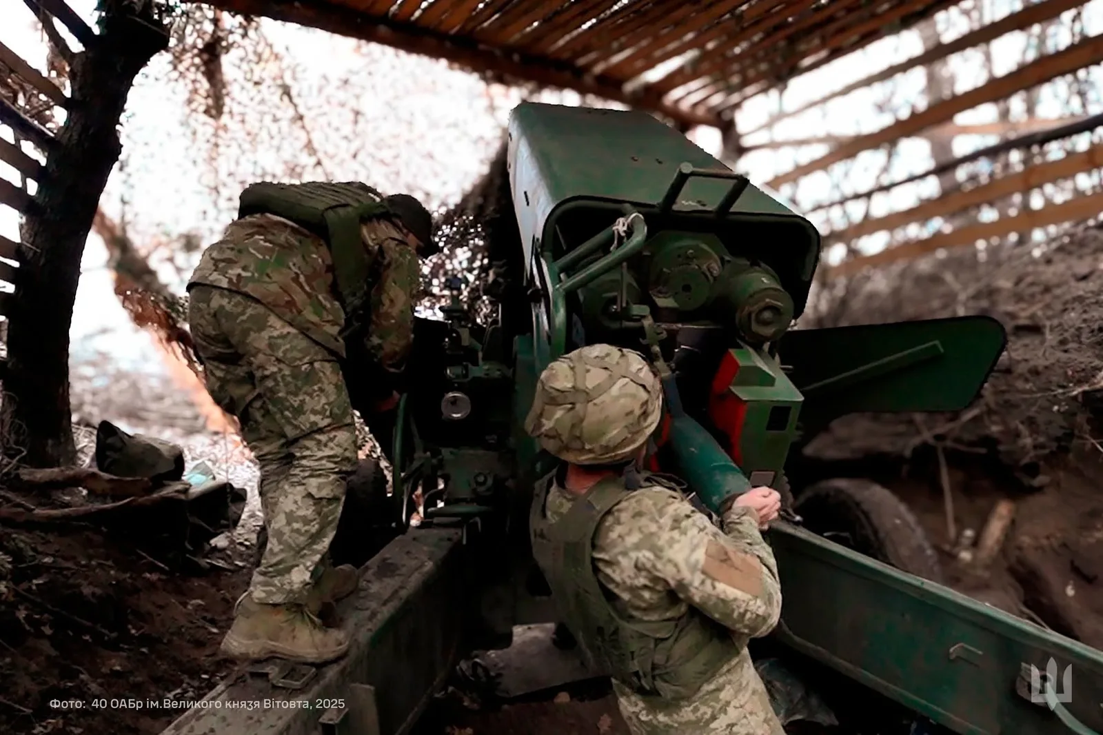 Двоє військових працюють біля артилерійської гармати в укритті. Один тримає снаряд біля механізму заряджання, другий нахилився з протилежного боку установки.