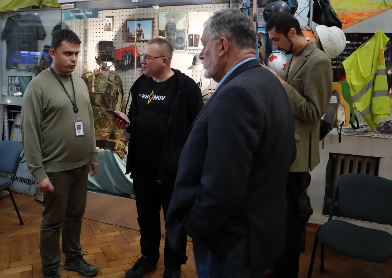 Чоловік в оливковому одязі розмовляє з чоловіком в окулярах, який тримає смартфон. Поруч стоїть сивий чоловік у темному піджаку. Чоловік праворуч у зеленуватому піджаку дивиться вниз на предмет у своїх руках.