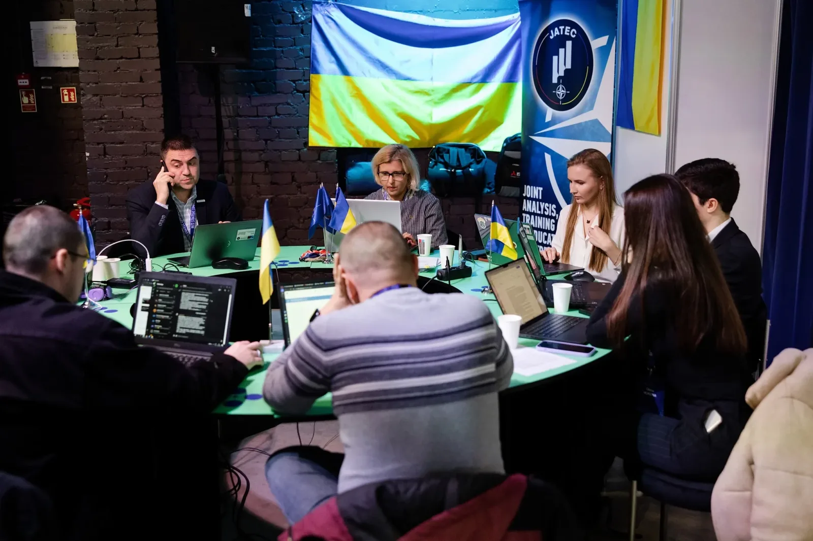 Група людей сидить за столами з відкритими ноутбуками та настільними прапорцями. Чоловік ліворуч тримає телефон біля вуха. Позаду висить український прапор і стоїть банер «JATEC».