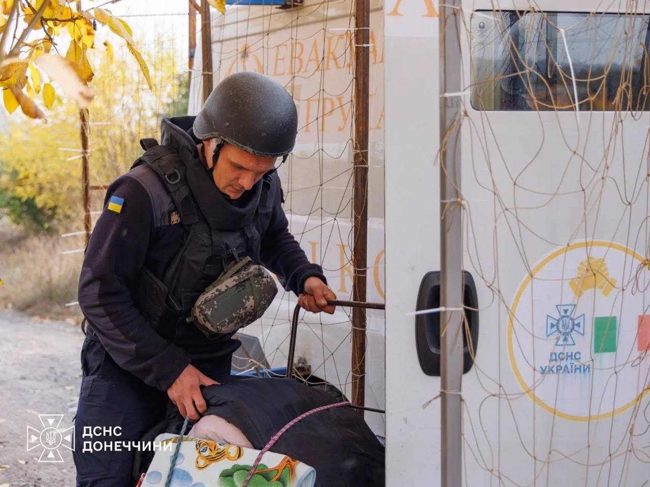 Рятувальник ДСНС у касці та бронежилеті допомагає людині сісти до евакуаційного автомобіля. На дверях машини видно символіку ДСНС України. Фото: ДСНС Донеччини