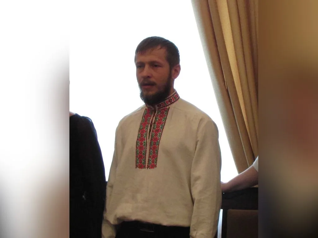 Чоловік із бородою у світлій вишиванці з червоно-зеленим орнаментом стоїть у приміщенні біля вікна з бежевими шторами.