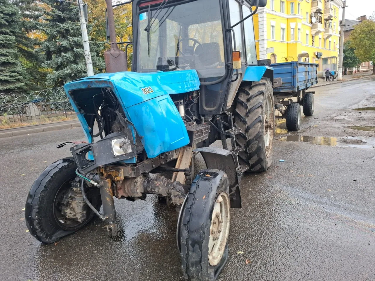Пошкоджений синій трактор із причепом стоїть на мокрій міській дорозі. У машини деформована передня частина, пошкоджені капот і переднє колесо.
